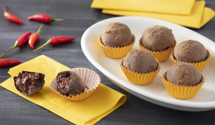 Fotografia em tons de amarelo de uma bancada cinza, sobre ela um prato oval com cinco trufas em forminhas amarelas, ao lado esquerdo um guardanapo amarelo com uma trufa cortada ao meio sobre ele. Ao fundo pimentas vermelhas.