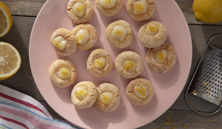 Foto da receita de biscoitos sem gluten e sem lactose com creme de limão servidos em diversas porções sobre um prato de porcelana rosa em cima de uma mesa de madeira com limões sicilianos em volta. Além disso, há um ralador e um pano