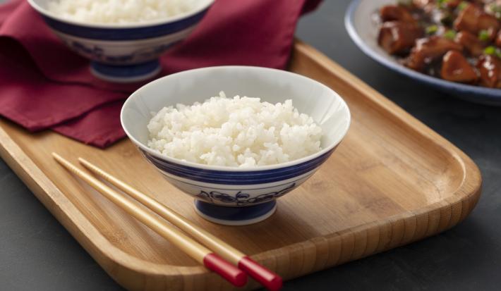 Fotografia em tons de marrom em uma bancada de madeira cinza escura com uma bandeja de madeira com um prato de cerâmica branca com detalhes em azul com o arroz japonês dentro dele. Ao lado, hashi e um pano na cor vinho.