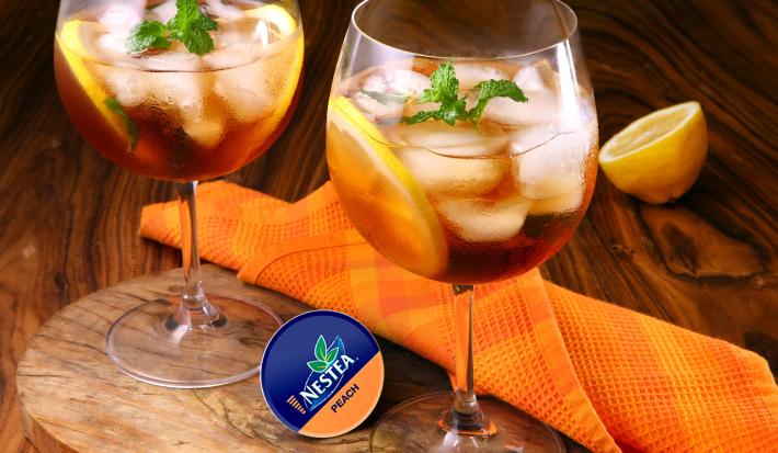 Fotografia em tons de laranja e azul em uma bancada de madeira clara, uma tábua de madeira, um pano laranja e duas taças de bebida com o Gin de Nestea Pêssego dentro delas.