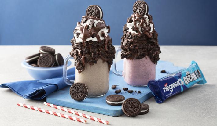 Fotografia em tons de azul, branco e marrom de uma bancada branca vista de frente, contém 2 copos transparentes um com milk-shake de baunilha e o outro de morango e ambos tem biscoitos de chocolates por cima para decorar. Ao lado o pacote do biscoito.
