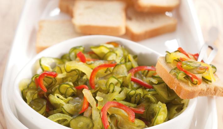 Fotografia em tons de marrom de uma bancada de madeira com um recipiente branco, sobre ele o relish de pepino e torradinhas.