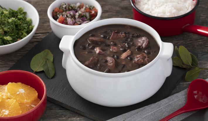 Fotografia em tons de vermelho em uma bancada de madeira escura, uma tábua preta com uma panela branca com a feijoada dentro dela. Ao lado, vários potinhos com laranja, couve, arroz e farofa.