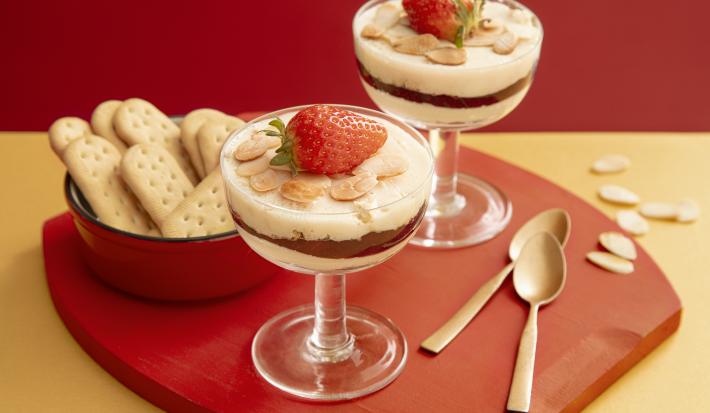 Foto da receita de Panacota de Frutas Vermelhas e Biscoito na taça. Observa-se duas taças com camadas e decoradas com lâminas de amêndoas e um morango por cima. Ao lado esquerdo, um potinho com biscoitos inteiros.