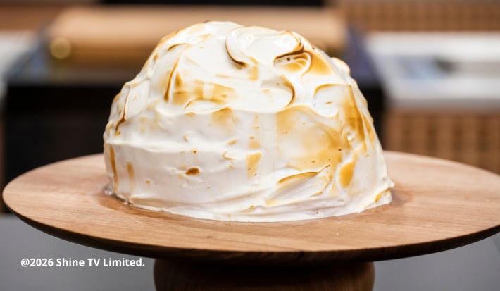 Sobremesa coberta com merengue tostado dourado sobre suporte de madeira, com fundo desfocado de cozinha.