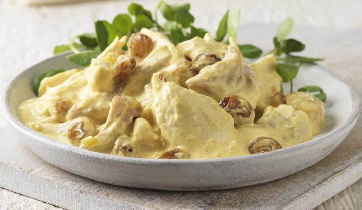 Fotografia em tons claros de uma salada de frango com molho ao curry e algumas folhas em um recipiente de cerâmica raso na cor branco. O prato está sobre uma peça de madeira em tom claro.