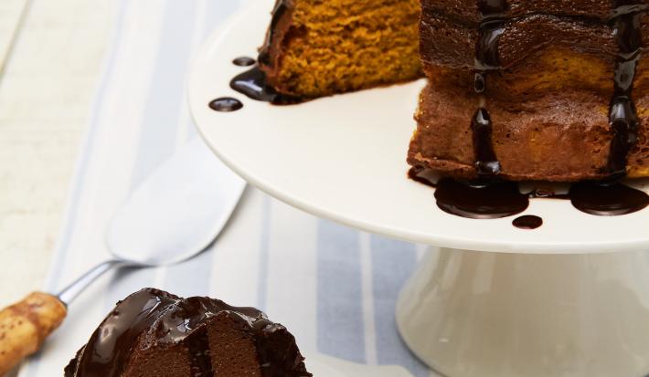 Fotografia em tons de marrom em uma bancada de madeira com um pano branco com listras azuis. Ao centro, um prato branco redondo raso com uma fatia do bolo de cenoura com chocolate. Ao fundo, um suporte branco para bolos com o bolo inteiro.