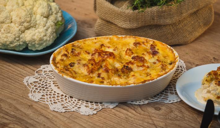Fotografia em tons de amarelo em uma bancada de madeira clara, um paninho de renda e um recipiente branco oval com a couve-flor gratinada feita na AirFryer Mondial dentro dele. Ao lado, um prato com couve-flor crua, um outro prato com uma fatia.