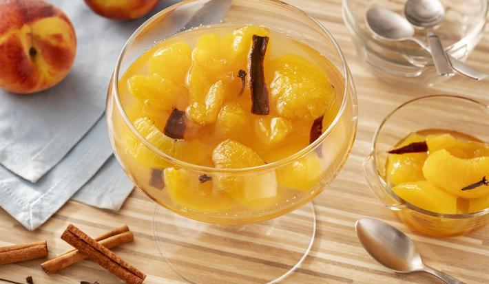 Fotografia em tons de amarelo em uma bancada de madeira de cor marrom. Ao centro, um recipiente de vidro contendo o doce de pêssego. Ao lado, há canelas em pau, um recipiente pequeno com doce e uma colher. Ao fundo, um pano azul claro com 2 pêssegos.
