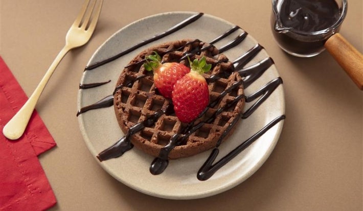 Fotografia de um prato de vidro com o waffle, dois morangos inteiros e a calda de nescau.
