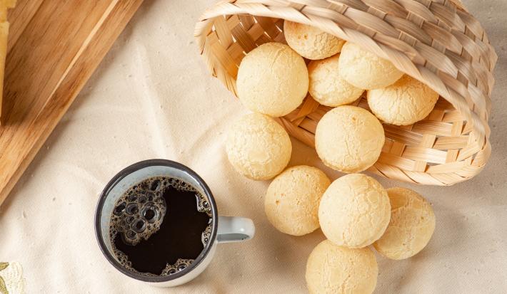 Foto da receita de Pão de Queijo MAGGI MEU SEGREDO 7 Vegetais. Observa-se os pães de queijo dispostos numa mesa saindo de uma cestinha de palha, acompanhados de uma xícara pequena de café vista de cima.
