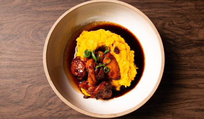 Fotografia vista de cima de uma receita com arroz cremoso na cor amarela sobre de um molho com shoyu. Por cima do arroz tem uns pedaços de frango caramelizados, e cebolinha para enfeitar.