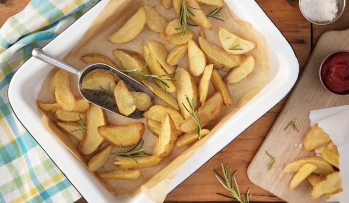 Imagem vista de cima de uma travessa retangular branca com batatas fritas rústicas, ramos de alecrim e um pegador. Ao lado, uma tábua com um saquinho de batatas, um potinho de ketchup e um potinho de sal.