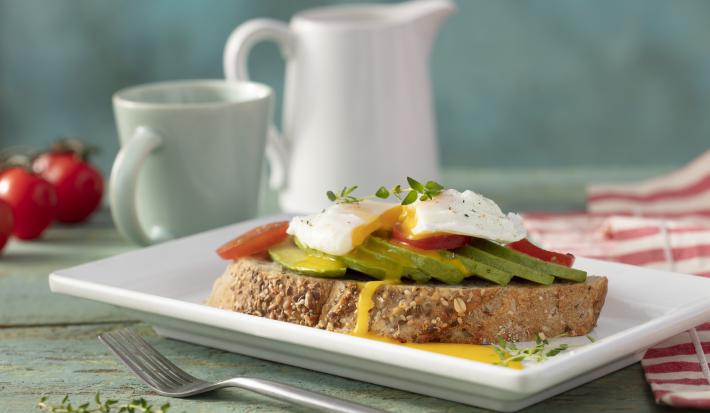 Fotografia em tons de vermelho em uma bancada de madeira verde clara, um paninho vermelho listrado, um prato quadrado branco com uma torrada e um ovo poché em cima com abacate. Ao fundo, tomates, xícara e jarra com café.