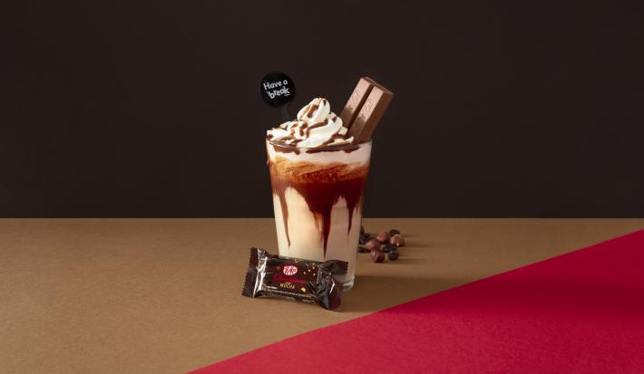 Foto da receita de Frapê Avelã. Observa-se Um fundo escuro marrom e ao centro um copo alto, decorado com calda de chocolate e dentro a bebida cremosa. Ao topo, chantilly e um KIT KAT Dark espetado. Avelãs decoram a parte direita do copo.