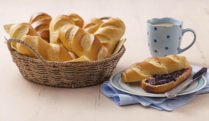 Fotografia em tons de azul em uma bancada de madeira clara, um pano azul, um prato branco redondo com detalhes em azul e um pão doce aberto com geleia no meio em cima dele. Ao lado, uma cesta de vime com vários pães e uma xícara azul com café.