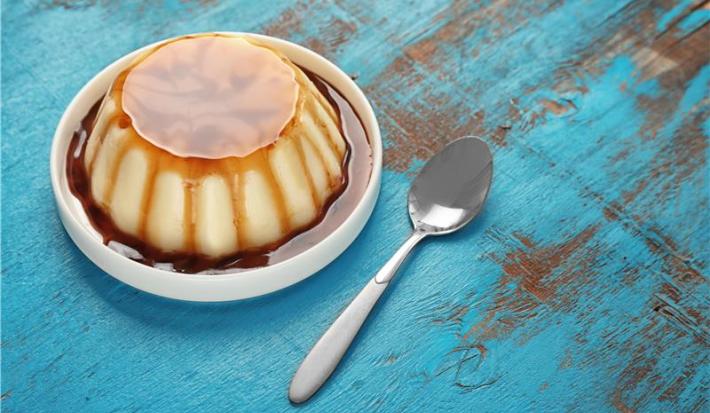 A imagem mostra um Pudim de Tapioca Simples cremoso, com uma textura suave e um caramelo dourado brilhante por cima. O pudim é servido em uma forma elegante, destacando sua cor branca e a consistência delicada.