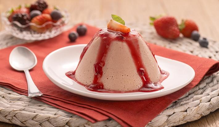 Foto em tons de vermelho da receita de flan de morango com calda de frutas vermelhas servido em um prato de porcelana branco sobre um pano vermelho com uma colher prateada ao lado, morangos e mirtilos ao fundo junto com uma taça de vidro com frutas