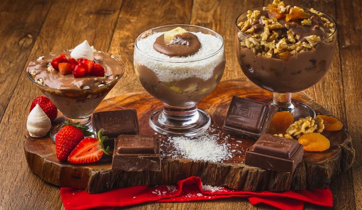 Foto da receita de Taças Cremosas, composta por três taças de vidro lado a lado sobre uma tábua de madeira com em um tecido vermelho. Cada uma está com um creme em tom marrom e alguns ingredientes compondo-as e a tábua está decorada.