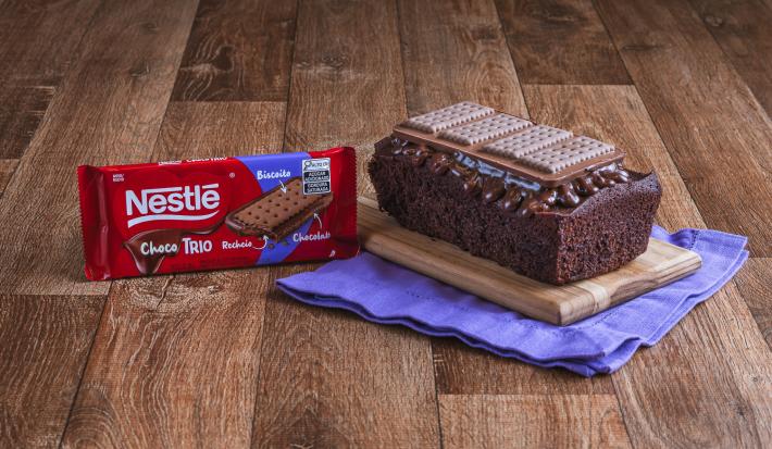 Foto da Receita de Bolo de Chocolate com Cobertura de Choco Trio. Observa-se um bolo em formato inglês sobre uma tábua de madeira e um pano roxo.