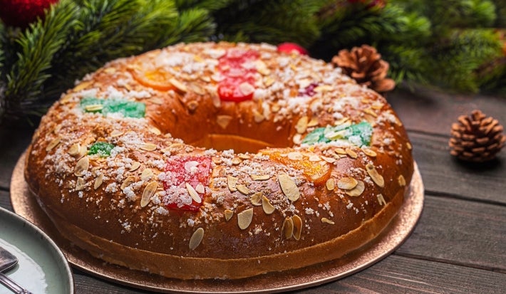 Fotografia de um bolo decorado com frutas cristalizadas vermelhas e verdes, amêndoas fatiadas e açúcar. Apresentado em um prato prateado sobre uma superfície de madeira escura, rodeado por ramos de pinheiro e enfeites natalinos.