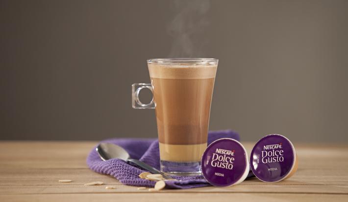 fotografia em tons de marrom, roxo e cinza de uma bancada de madeira marrom vista de frente, contém um pano roxo com uma colher, uma xicara transparente com café e duas capsulas roxa de café