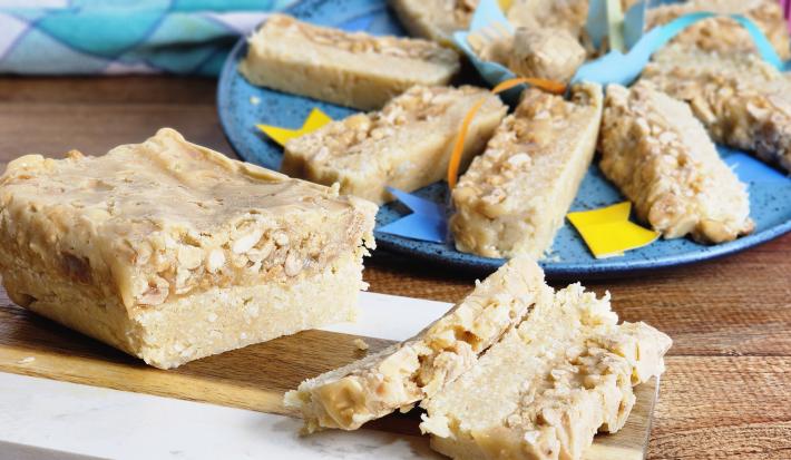 Fotografia em tons de azul com um prato azul ao centro. Em cima do prato existe vários pedaços de um doce retangular de cor caramelo feito com amendoim e coco ralado.