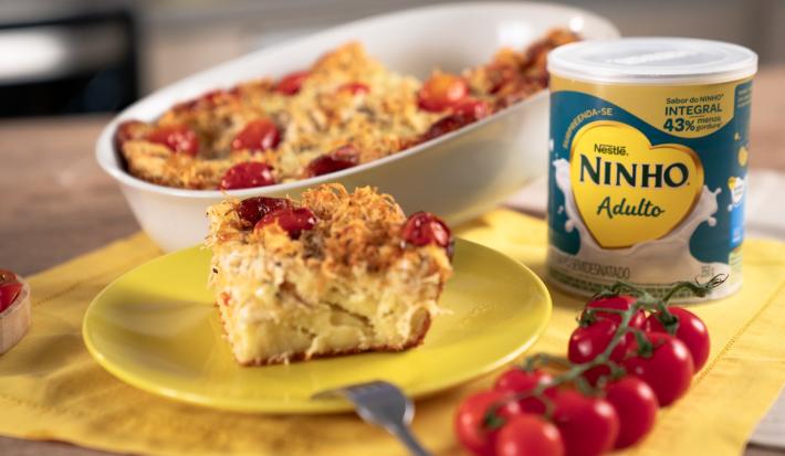 Fotografia em tons de amarelo em uma bancada de madeira, um pano amarelo e um prato amarelo redondo ao centro com um pedaço de torta de frango em cima. Ao fundo um  recipiente com a torta inteira e a lata de Ninho Adulto ao lado.