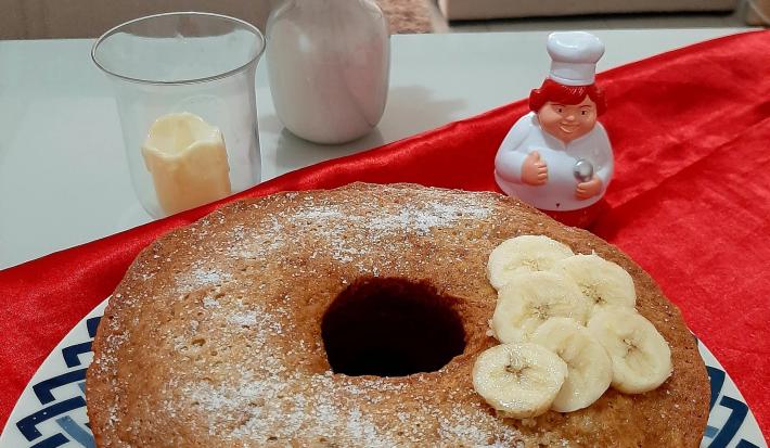 Fotografia em tons de azul em uma mesa com um pano vermelho, um prato redondo raso com desenho em azul e um bolo de banana com canela e rodelas de banana em cima dele.