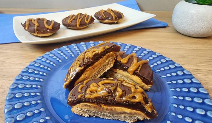 Fotografia em tons de azul em uma bancada de madeira e um prato azul redondo com tortinhas de caramelo salgado com chocolate e aveia em cima dele.