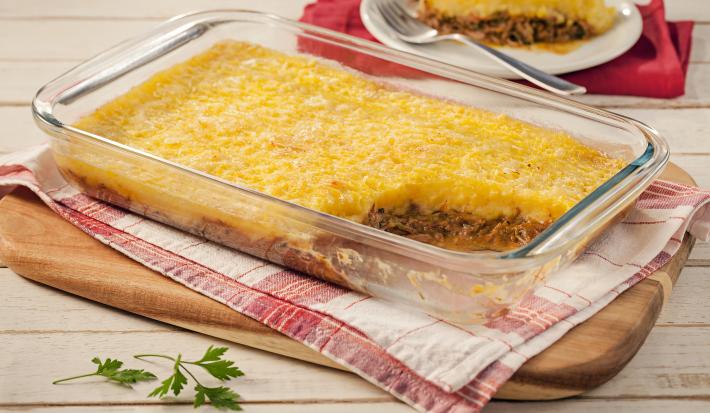 Imagem de uma travessa retangular transparente com a receita de escondidinho com carne desfiada sem um pedaço, mostrando a camada de carne e a camada de purê. Ao fundo, um prato branco com o pedaço e um garfo.