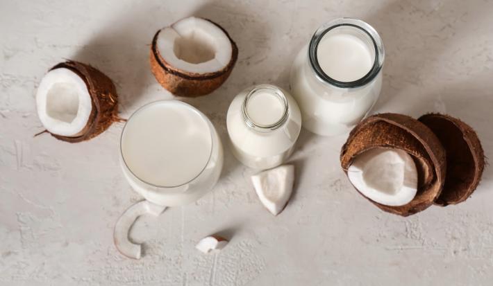 Fotografia vista de cima de duas garrafas decorativas de vidro, uma jarra branca e um copo grande e largo. Todos esses recipientes têm bebida vegetal de coco. Ao lado tem cascas, pedaços e polpa do coco pela metade sobre uma mesa cinza clara.