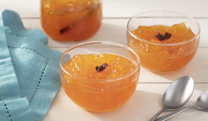 Fotografia em tons de laranja em uma bancada de madeira branca, um pano azul, três potinhos de vidro com o doce de mamão ralado e em pedaços pequenos com cravos. Ao lado, duas colheres.