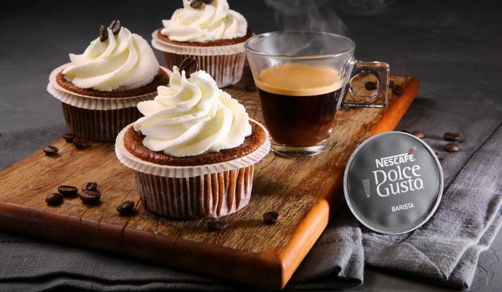 Fotografia em tons de marrom em uma bancada cinza com pano cinza escuro, uma tábua de madeira com três forminhas com cupcakes feitos com café e leite dentro delas. Ao lado, uma xícara de café e a cápsula do Café Barista Dolce Gusto.