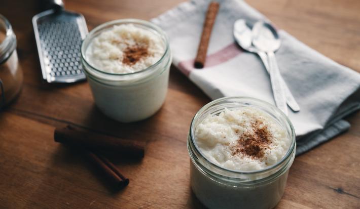 Fotografia de dois potes pequenos de vidro com arroz doce e canela polvilhada por cima. No fundo da foto, duas colheres de sopa e uma canela em pau sobre um pano de pano branco com uma listra vermelha. Ao lado dos potes, tem duas canelas em pau.
