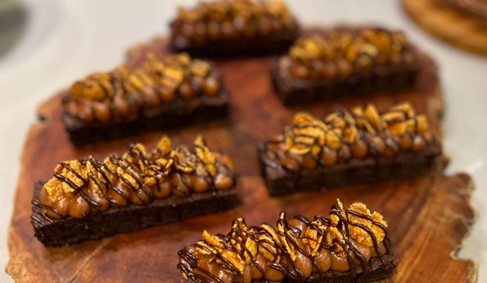 Fotografia em tons de marrom com uma bancada de madeira ao centro. Em cima da bancada existe 6 sobremesas com base de brownie e cobertas com um creme de caramelo e chocolate