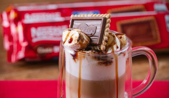 Fotografia em tons de vermelho com uma caneca de vidro ao centro. Dentro da caneca existe uma receita de café com leite finalizada com marshmallow e Choco Biscuit NESTLÉ