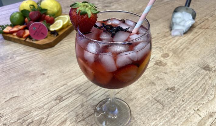 Fotografia em tons de rosa, em uma bancada de madeira clara, uma tábua de fundo com pitaya, limão, morango e cápsula de Dolce Gusto, e uma pá com gelo. Ao centro, uma taça com gelo e uma bebida rosa dentro com um morango decorando a taça.