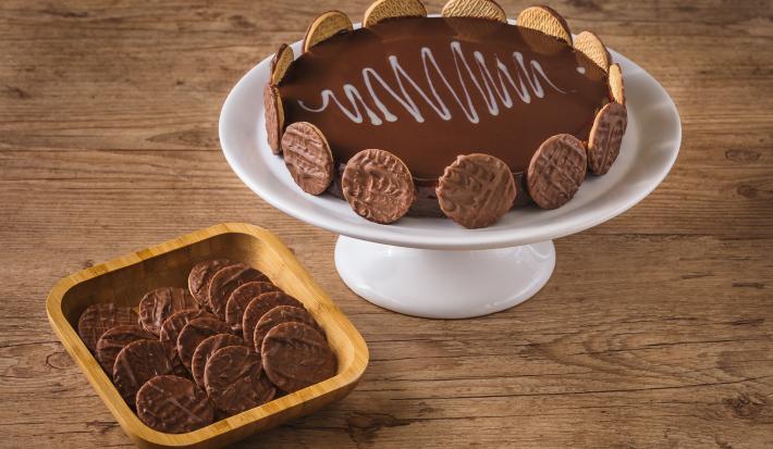 Imagem de uma torta decorada com biscoitos ao redor, disposta num prato branco alto, ao lado de uma tigela de madeira com mais biscoitos dentro