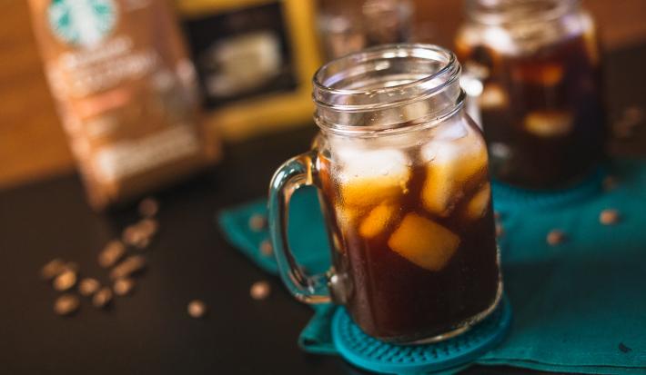 Fotografia em tons de preto de uma bancada preta, com um paninho verde e dois copos com alça com gelo e café.