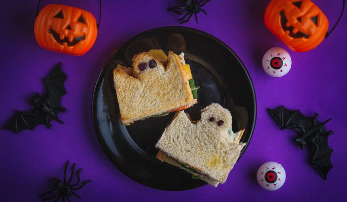 Imagem de um ângulo de cima. Numa mesa em tom roxo escuro há um prato preto com dois sanduíches de pão de forma cortados em formato de fantasmas, com olhos pretos. Ao redor há abóboras, morcegos e aranhas de mentira.