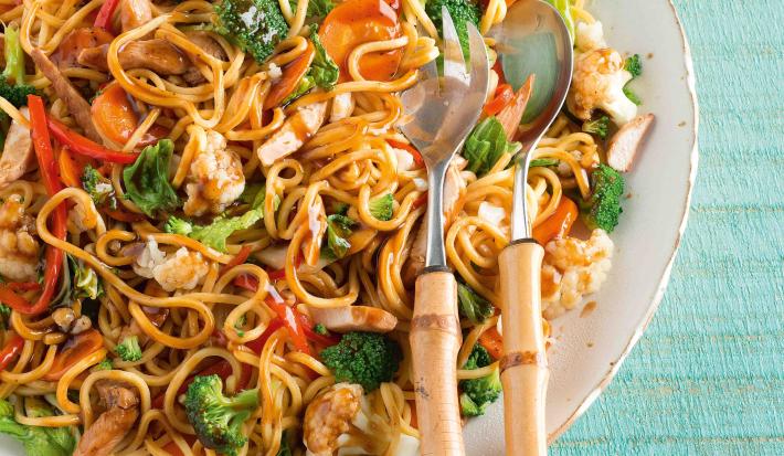 Foto de um prato branco cheio de yakissoba com frango e legumes. Encostado ao prato há duas colheres