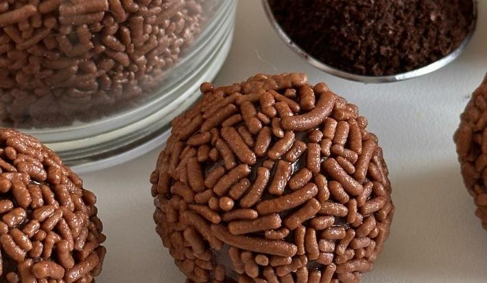 Fotografia em tons de branco com vários brigadeiros ao centro. Ao lado dos brigadeiros existe uma colher de café em pó.