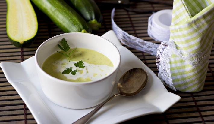 Fotografia de um recipiente fundo branco de vidro com uma sopa de abobrinha e cará, com um creme branco por cima e salsinha. A sopa está em cima de um prato de vidro branco e quadrado, com uma colher de sopa apoiada nele, sobre uma mesa de madeira.