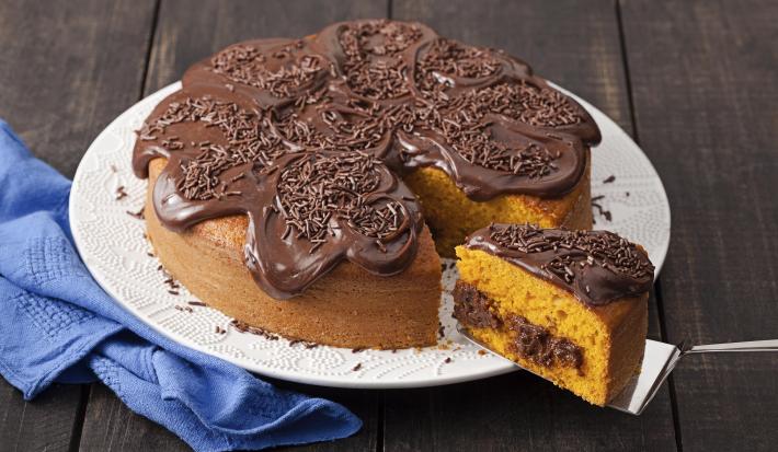 Fotografia em tons de marrom, azul e branco de uma bancada de madeira com um paninho azul e um prato redondo branco, sobre ele um bolo de cenoura com recheio e cobertura de chocolate .