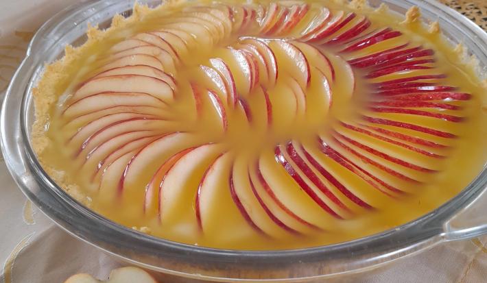 Foto da receita de Torta de Maça Dona Kilza. Observa-se um recipiente refratário transparente redondo e grande com a torta. A cobertura é feita de maçãs laminadas finas dispostas em círculos com um creme brilhoso de laranja.