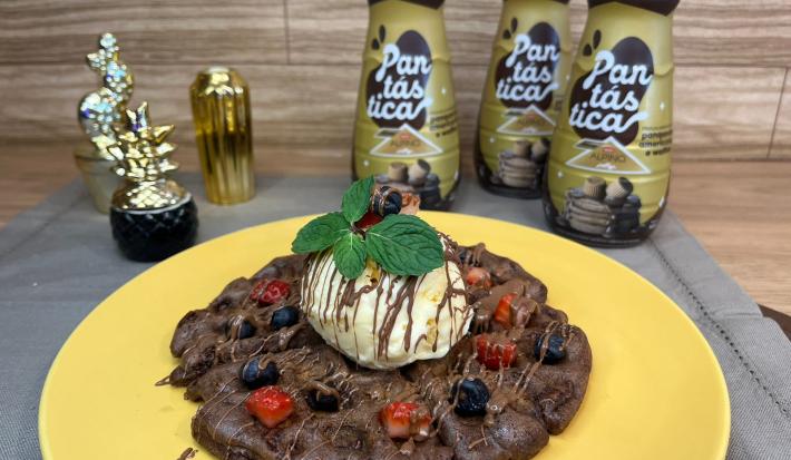 Fotografia em tons de dourado com um prato amarelo ao centro. Em cima do prato existe um waffle de chocolate com cobertura de chocolate derretido, frutas vermelhas e sorvete de creme. Em cima do sorvete existe folhas de hortelã.