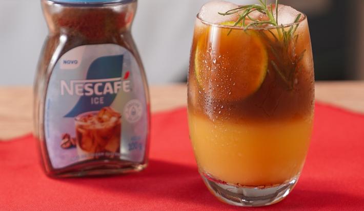 Fotografia de um copo de vidro com uma bebida de café gelado com tônica e laranja. A bebida está decorada com alecrim, e está ao lado de uma embalagem de nescafé gelado, sobre um pano vermelho.