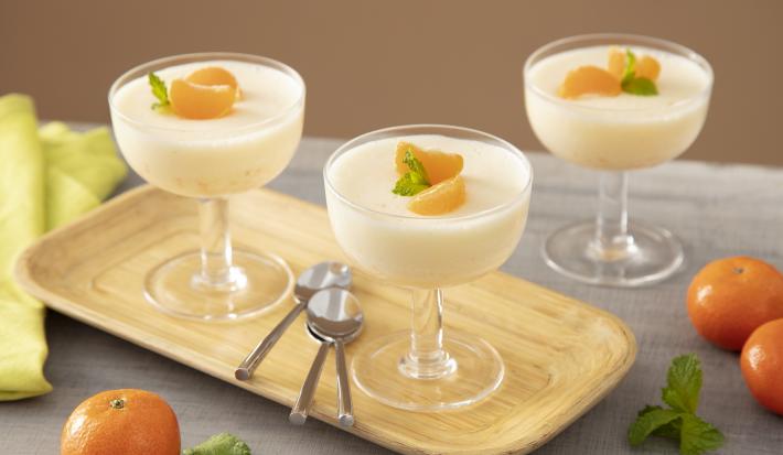 Foto da Receita de Mousse de Tangerina. Observa-se três taças de vidro com a mousse dispostas em uma tábua de madeira. Tangerinas, folhas e três colheres de sobremesa também decoram a foto