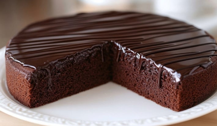 Em cima de uma mesa, prato redondo com bolo de chocolate redondo e cobertura de chocolate cremoso.